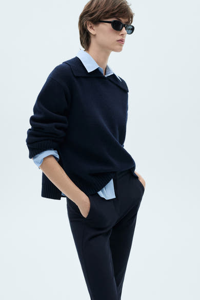 Donkerblauwe broek van MANGO, gedragen met een marineblauwe trui en een lichtblauwe blouse, handen in de zakken.