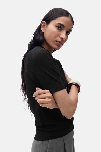 Zwarte rondehals trui van MANGO, gedragen met dikke armbanden en een grijze broek.