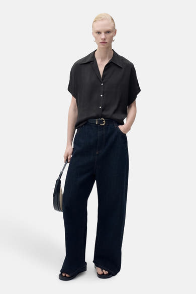 Chemisier noir à manches courtes par MANGO, porté avec un jean taille haute, une ceinture et un sac à main.
