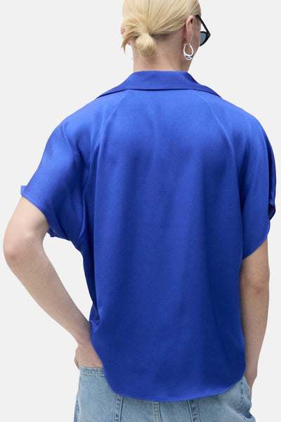 Chemise à manches courtes bleue de MANGO, vue de dos, avec un jean clair et des boucles d'oreilles argentées.
