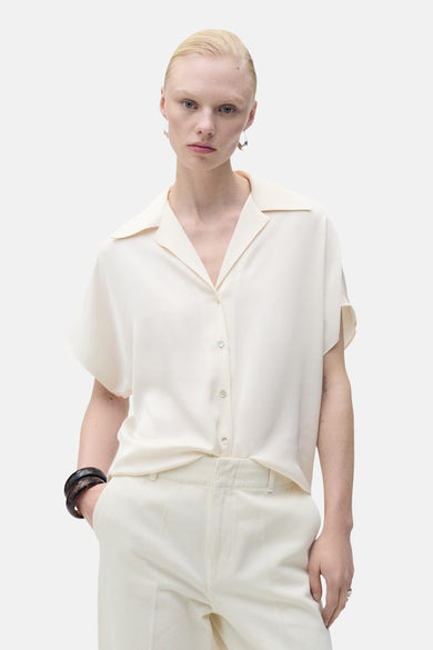 Chemise à manches courtes écru de MANGO, avec un col ouvert et des boutons, portée avec un pantalon blanc assorti et des bracelets foncés.
