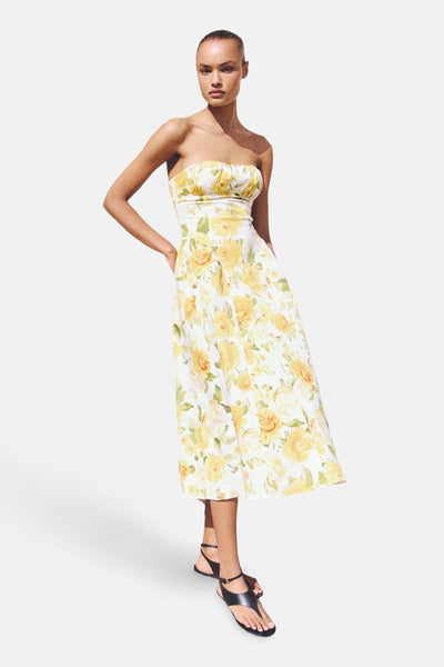 Robe jaune fleurie de MANGO, mi-longue, sans bretelles, portée avec des sandales noires à brides.
