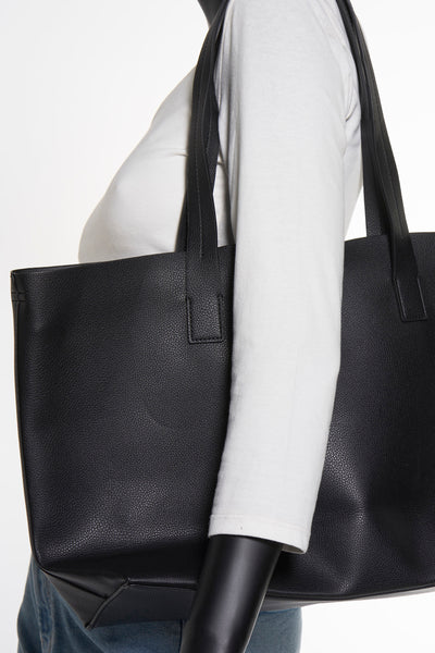 Sac à bandoulière noir en similicuir par MANGO, porté à l'épaule, sur mannequin avec chemise blanche et jean.
