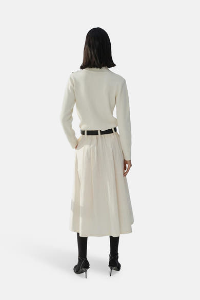 Pull à col rond écru de MANGO, porté avec une jupe midi taille haute, des collants noirs et des talons.