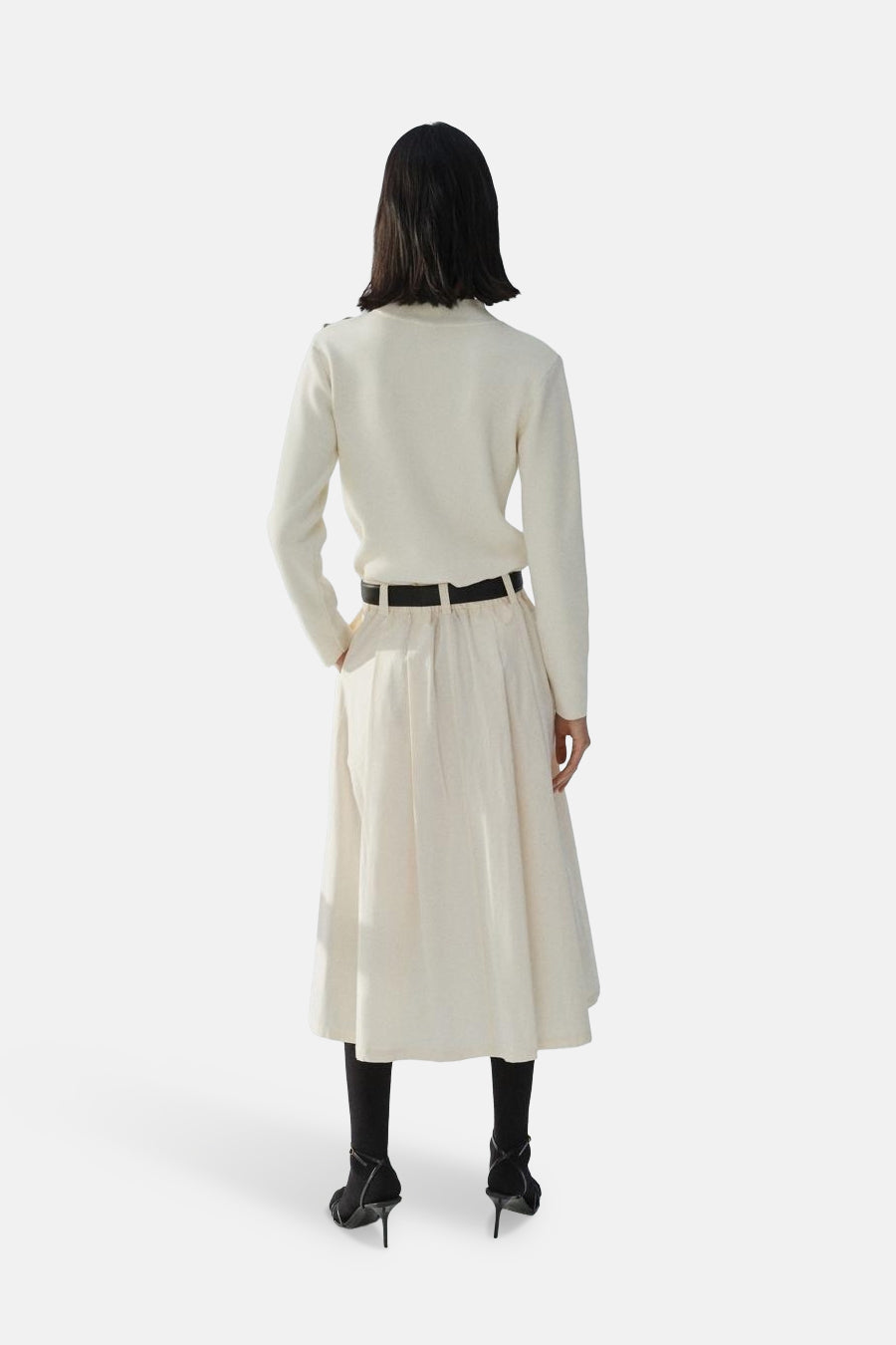 Pull à col rond écru de MANGO, porté avec une jupe midi taille haute, des collants noirs et des talons.