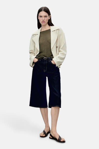 Pull à col rond kahki de MANGO, porté avec un pantalon court large en denim bleu et un manteau beige.
