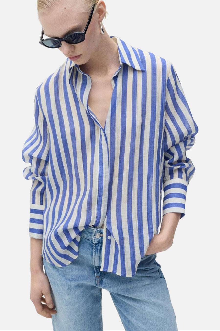 Chemise à manches longues bleue et blanche rayée de MANGO, portée avec un jean bleu clair et des lunettes de soleil noires.
