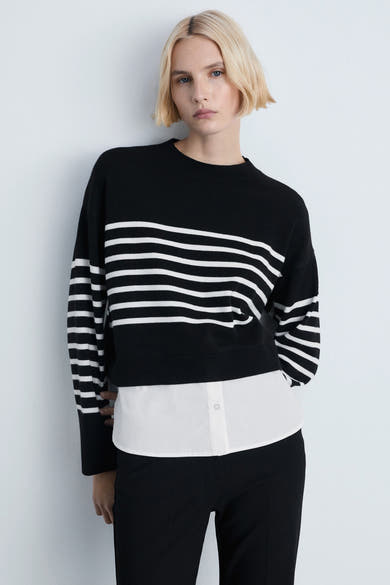 Zwarte rondehals trui van MANGO, met horizontale witte strepen, gedragen over een wit overhemd en een zwarte broek.
