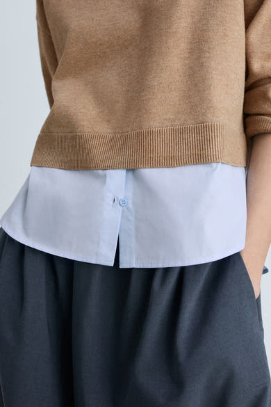 Bruine ronde trui van MANGO, gedragen over een lichtblauw overhemd en een grijze wijde broek.