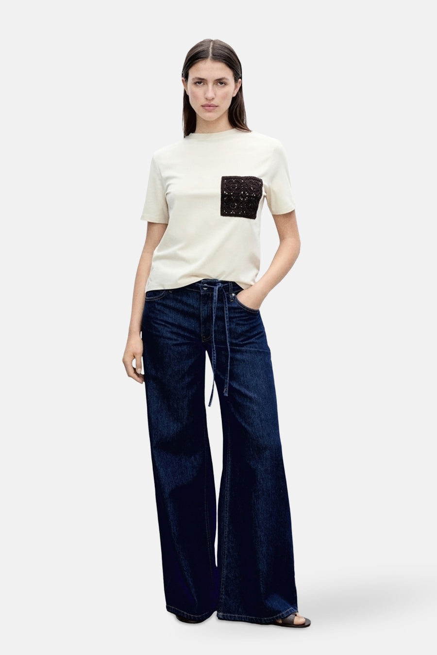T-shirt beige à manches courtes de MANGO, avec poche noire en dentelle, porté avec un pantalon taille haute.