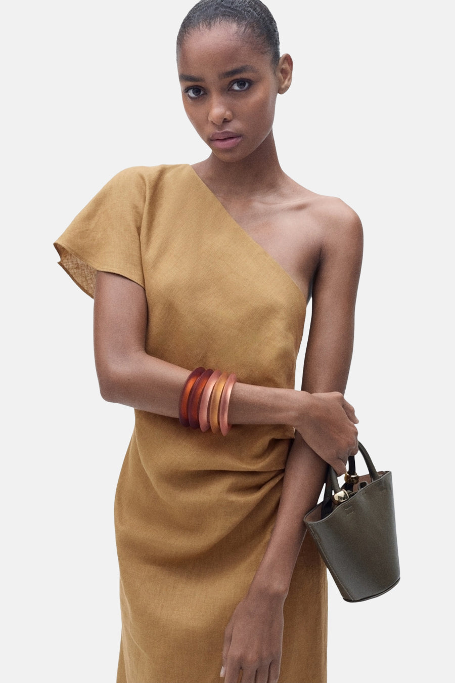 Bruine jurk van Mango, asymmetrisch met één schouder, geaccessoriseerd met armbanden en een handtas.