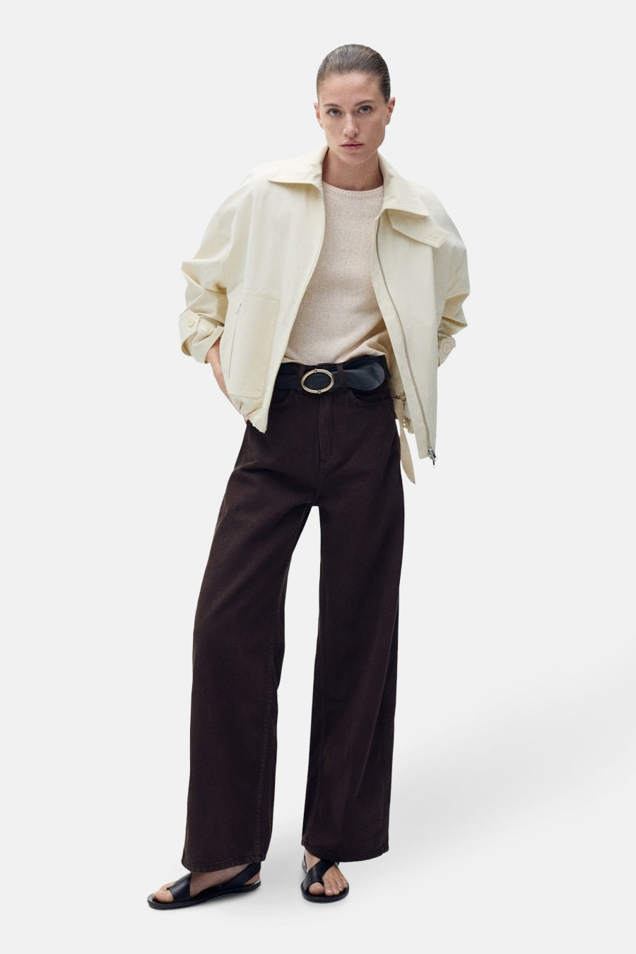 Pull à col rond beige de MANGO, porté avec une veste crème, un pantalon marron taille haute et une ceinture noire.
