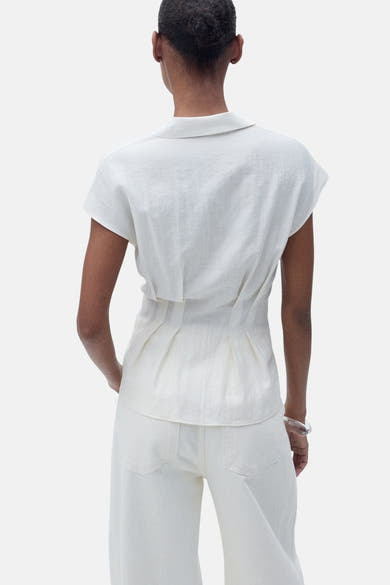 Chemisier écru à manches courtes de MANGO, détail plissé à la taille, porté avec un pantalon blanc ample.
