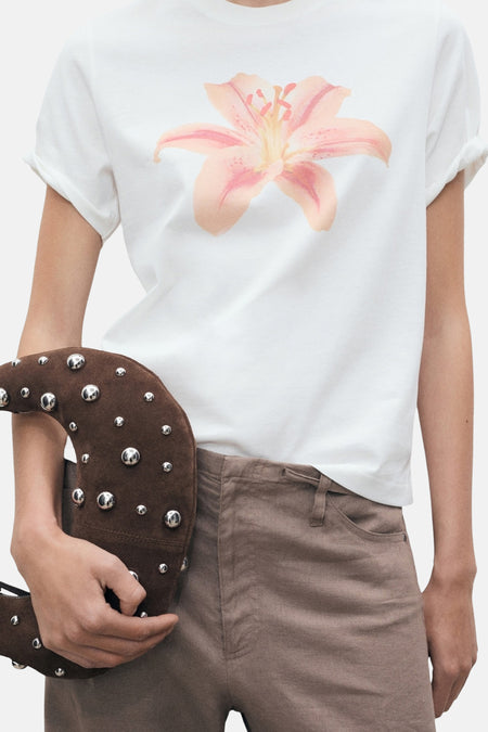 T-shirt à manches courtes blanc de MANGO avec fleur rose imprimée, porté avec un pantalon marron et un sac à main clouté.