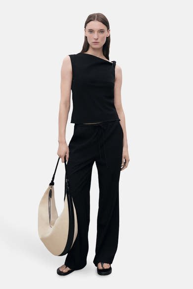 Zwarte tanktop van MANGO, met asymmetrische geknoopte halslijn, gedragen met een zwarte broek en een beige tas.
