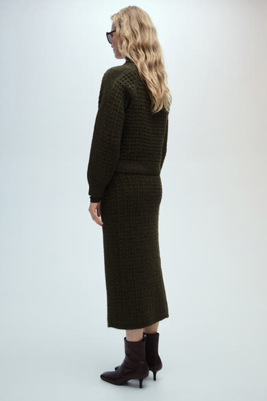 Pull col rond brun de MANGO, porté avec une jupe longue texturée et des bottines en cuir.