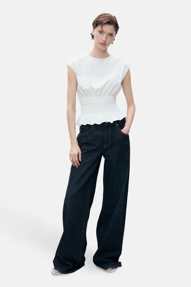 Wit mouwloos topje van MANGO, met smocktaille en volant, gedragen met een wijde jeans.