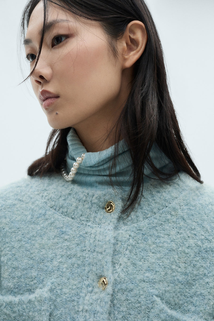 Blauwe MANGO-gilet met gouden knopen, gedragen met een parelketting en een blauwe coltrui.