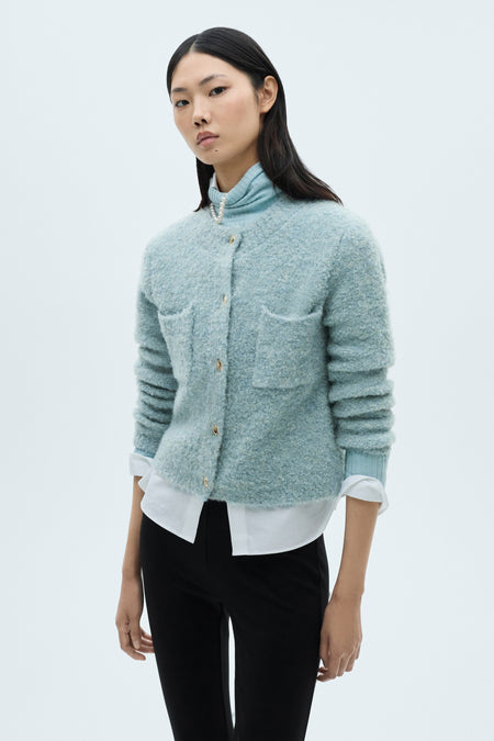 Blauwe gebreide bodywarmer van MANGO met opgestikte zakken en gouden knopen, gedragen over een wit overhemd en een zwarte broek.