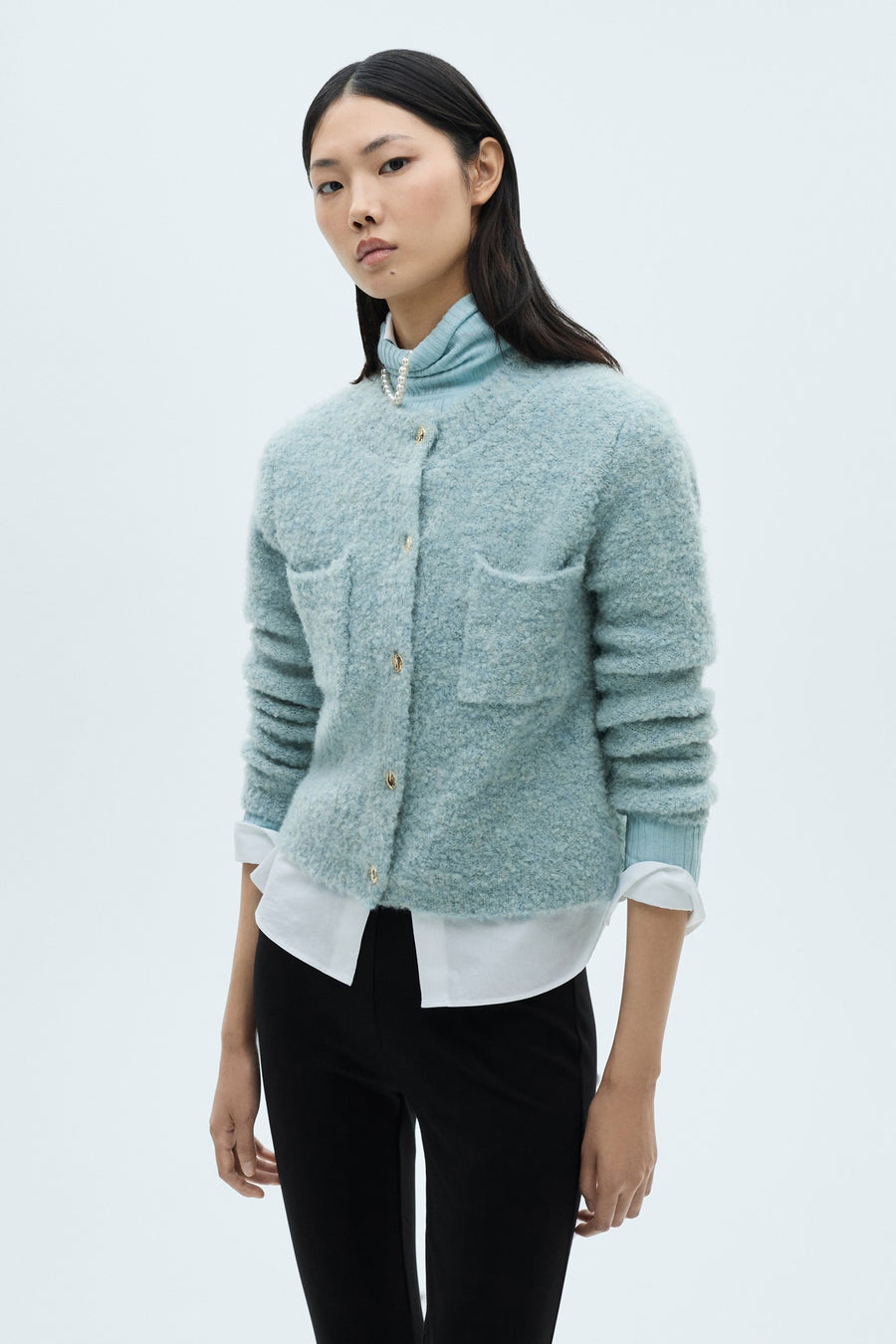 Blauwe gebreide bodywarmer van MANGO met opgestikte zakken en gouden knopen, gedragen over een wit overhemd en een zwarte broek.