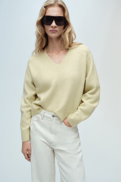 Groene V-hals trui van MANGO, gedragen met een witte wijde broek, achtergezicht.