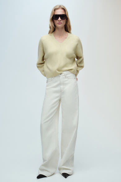 Groene V-hals trui van MANGO, gedragen met een gebroken witte wijde jeans en een zwarte zonnebril.