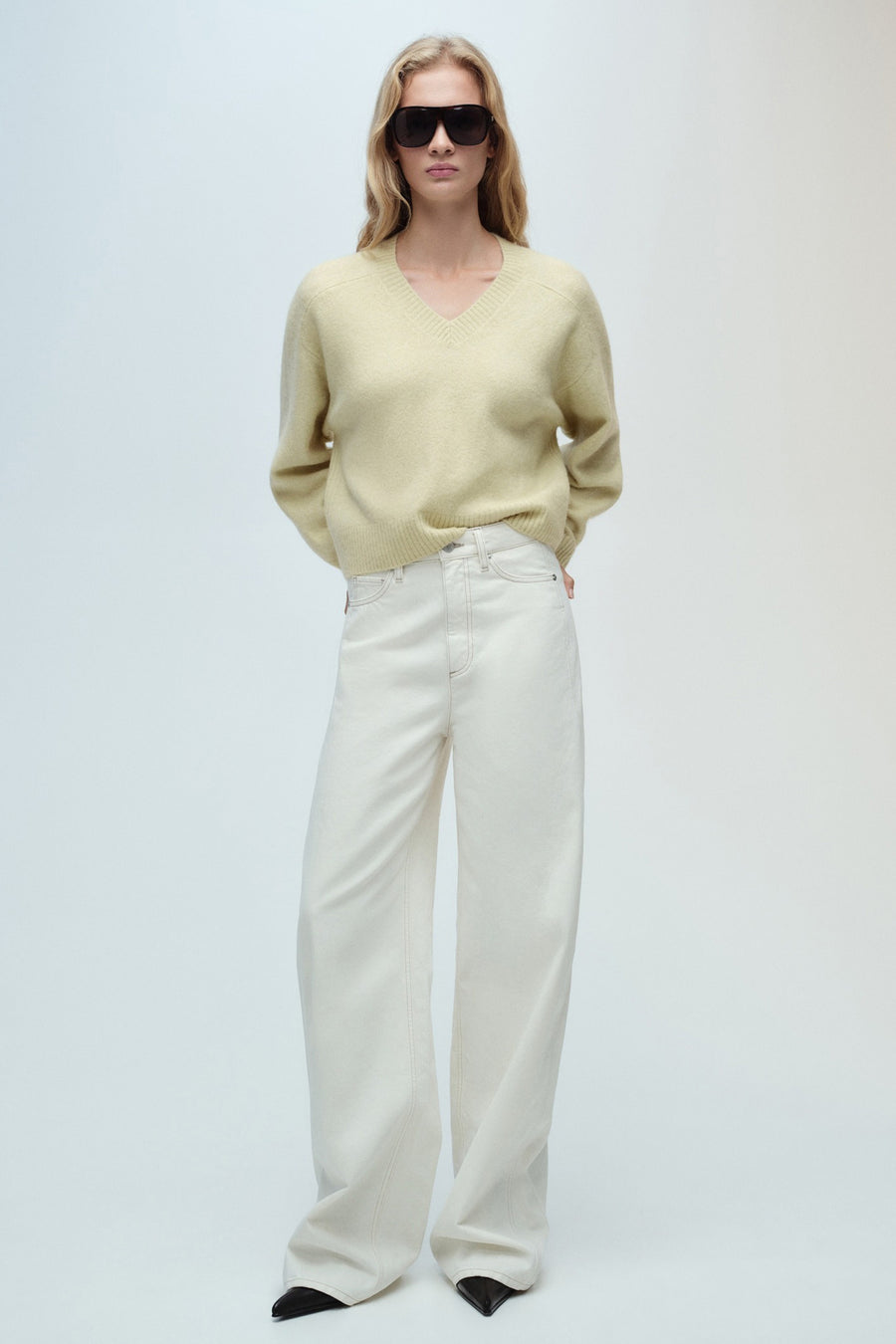 Groene V-hals trui van MANGO, gedragen met een gebroken witte wijde jeans en een zwarte zonnebril.