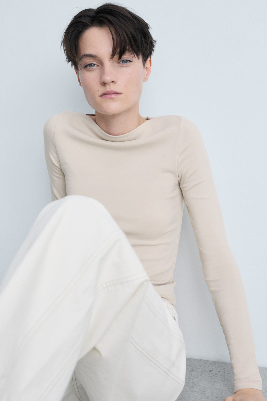 T-shirt beige à manches longues de MANGO, porté avec un pantalon blanc cassé, modèle assis contre un mur uni.
