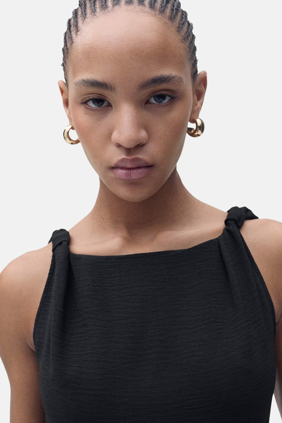 Robe noire de MANGO avec des bretelles nouées, affichant une texture subtile et des boucles d'oreilles dorées.