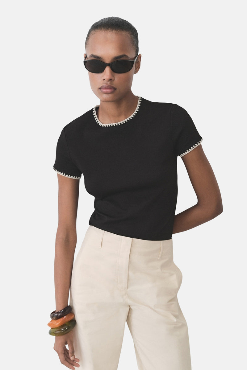 T-shirt noir à manches courtes de MANGO, bordure blanche festonnée au col et aux manches, porté avec un pantalon beige.
