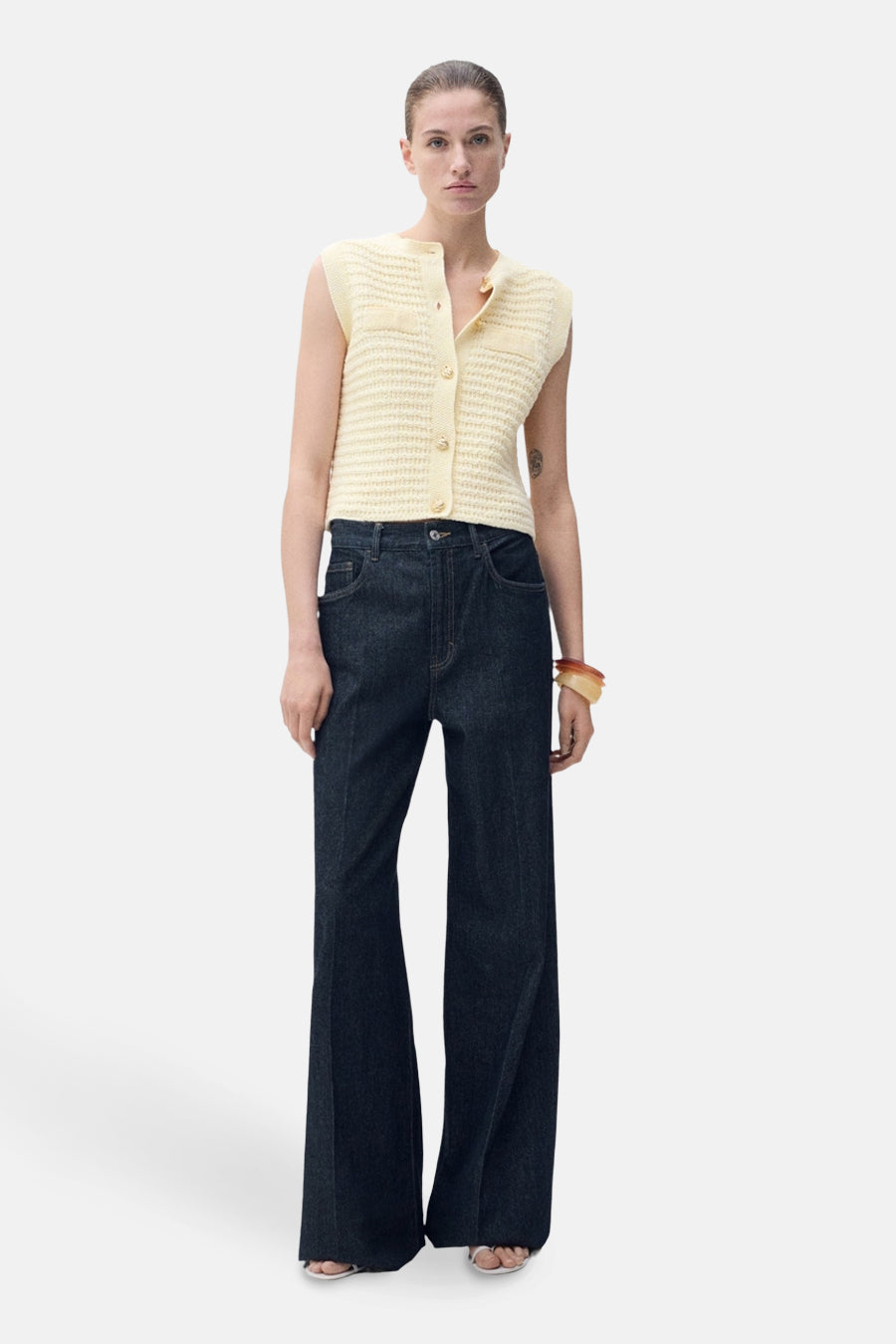 Gilet jaune de MANGO, boutonné et tricoté, porté avec un jean évasé et un bracelet.
