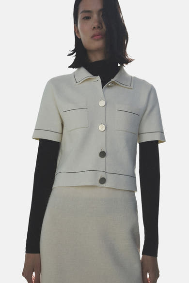 Ecru gilet van MANGO, met korte mouwen en contrasterende stiksels, gedragen over een zwarte top met lange mouwen.