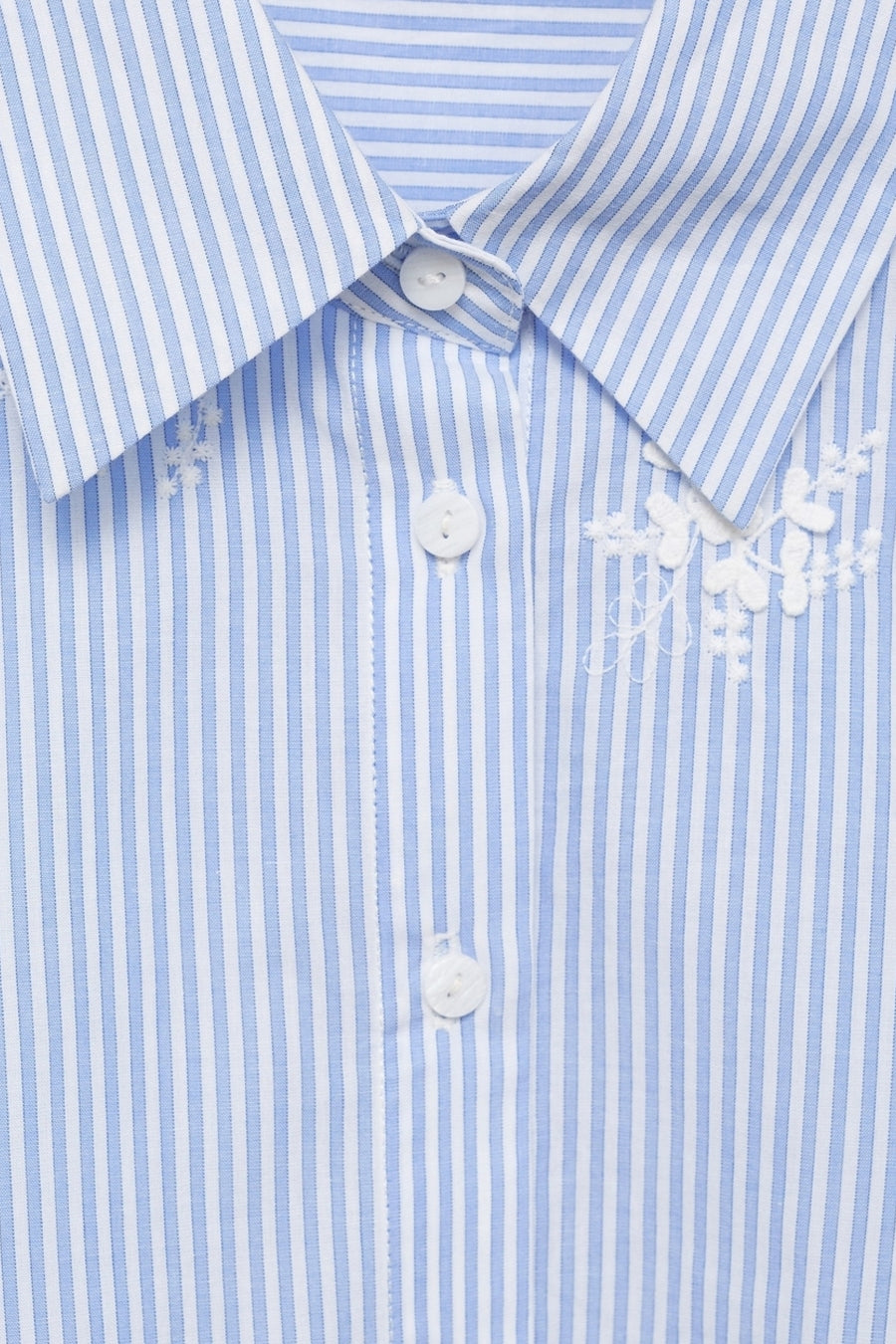 Chemise à manches longues bleue rayée de MANGO, avec broderie florale blanche sur le col et la patte de boutonnage.
