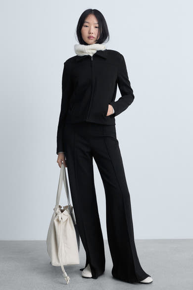 Zwarte broek van MANGO, uitlopend model met split onderaan, gedragen met een jasje en een beige tas.