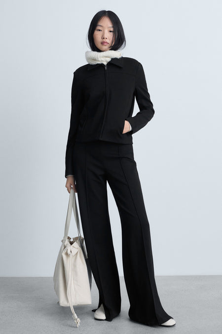Zwarte broek van MANGO, uitlopend model met split onderaan, gedragen met een jasje en een beige tas.