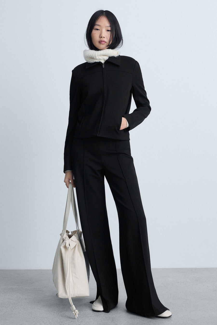 Zwarte broek van MANGO, uitlopend model met split onderaan, gedragen met een jasje en een beige tas.