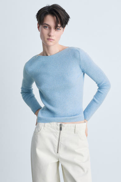 Pull col rond bleu de MANGO, en maille côtelée, porté avec un pantalon blanc à fermeture éclair.
