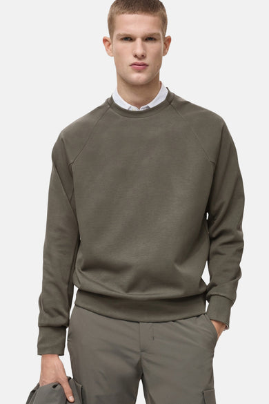 Sweat-shirt kaki par MANGO, porté sur une chemise blanche, avec un pantalon de la même couleur.