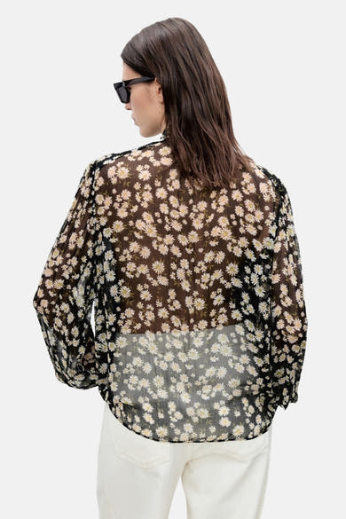 Chemisier noir à manches longues de MANGO, motif marguerites sur tissu transparent, porté avec un pantalon blanc.