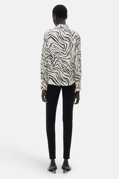 Chemisier à manches longues blanc à motif zébré, de MANGO, porté avec un pantalon noir.
