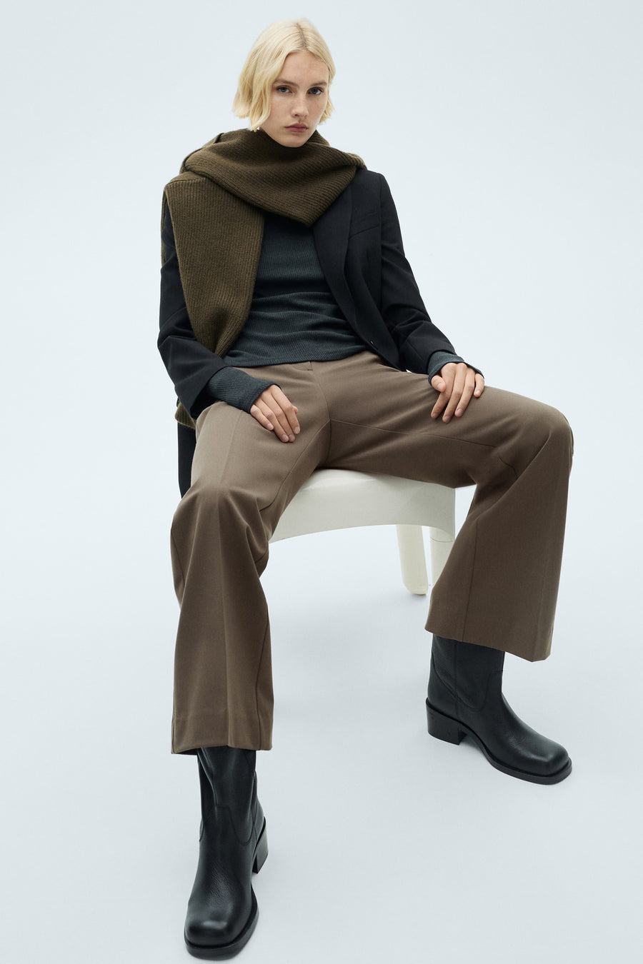 Pantalon de costume brun de MANGO, porté avec un pull gris, un blazer noir et des bottes en cuir.
