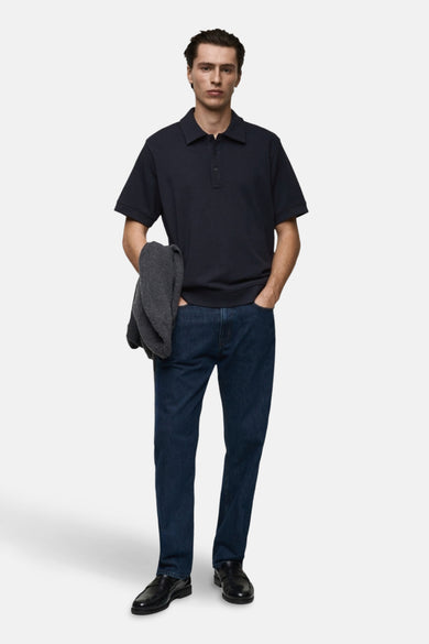Donkerblauwe polo van MANGO, gedragen met een blauwe spijkerbroek en zwarte schoenen, met een grijze trui over de arm.