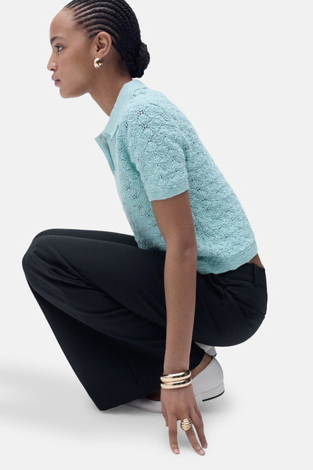 Blauwe polo van MANGO met ajourbreisel, gedragen met een wijde zwarte broek en gouden sieraden.