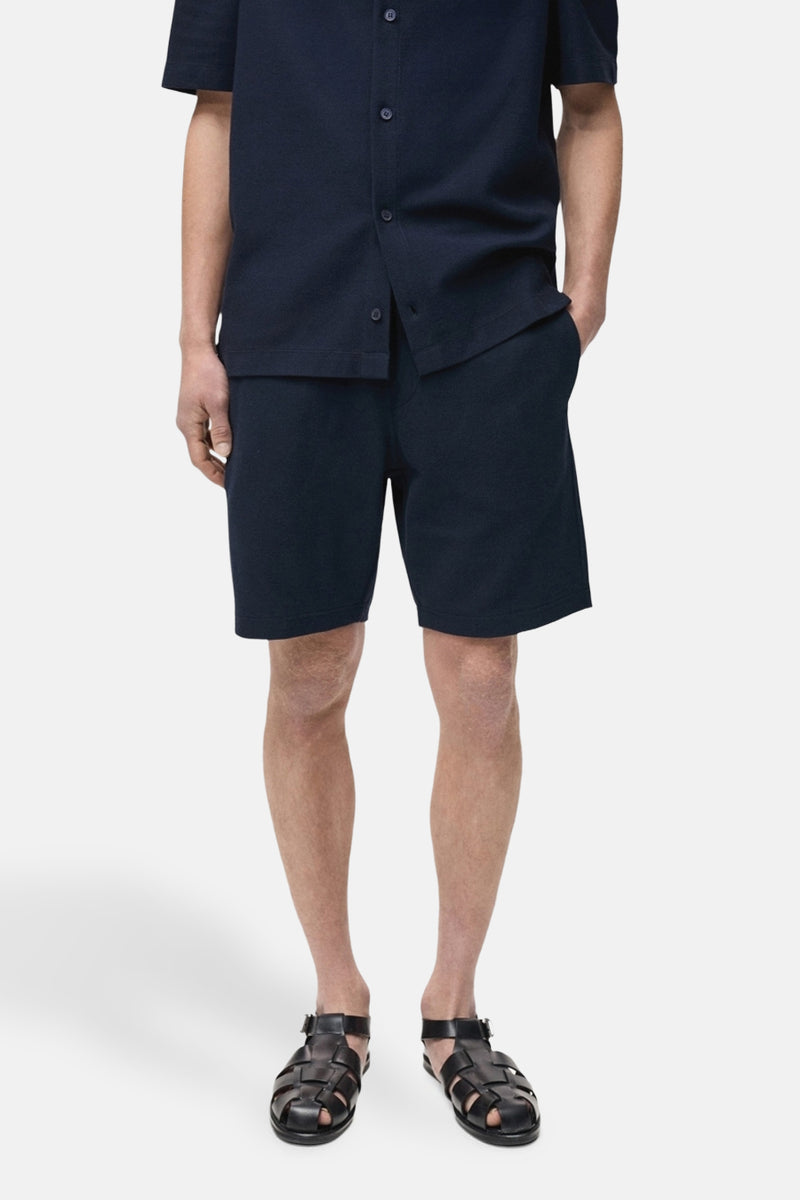 Short bleu foncé de MANGO, porté avec une chemise assortie et des sandales en cuir noir.

