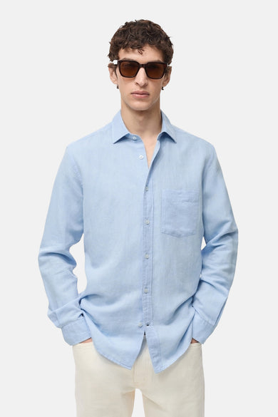Chemise à manches longues bleue de MANGO, avec poche poitrine et boutons clairs, portée avec un pantalon écru.