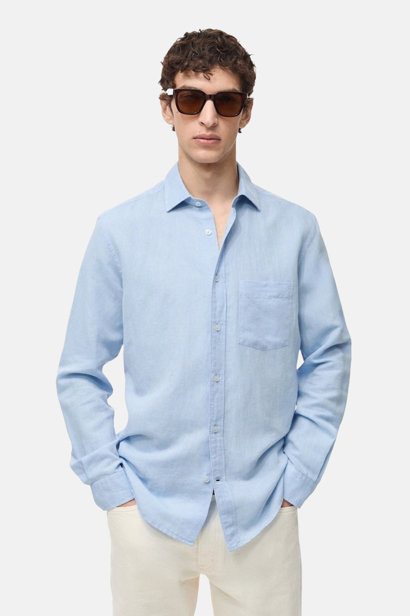 Chemise à manches longues bleue de MANGO, avec poche poitrine et boutons clairs, portée avec un pantalon écru.