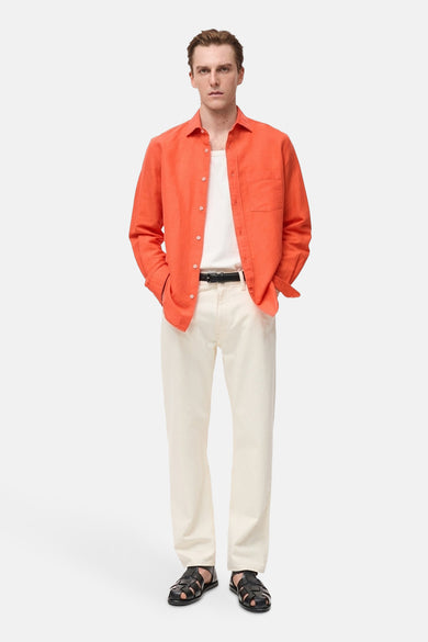 Chemise orange à manches longues de MANGO, portée ouverte sur un t-shirt blanc, avec un pantalon blanc cassé et des sandales en cuir noir.