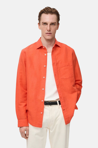 Chemise orange à manches longues de MANGO, portée sur un t-shirt blanc avec un pantalon clair et une ceinture noire.
