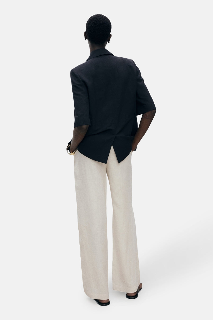 Pantalon beige de MANGO, coupe ample, en lin, porté avec un blazer noir, vue de dos.