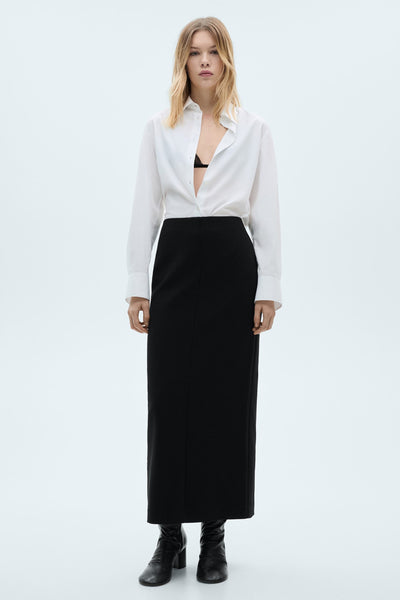 Chemise à manches longues blanche de MANGO, portée ouverte sur un soutien-gorge noir et avec une jupe noire.
